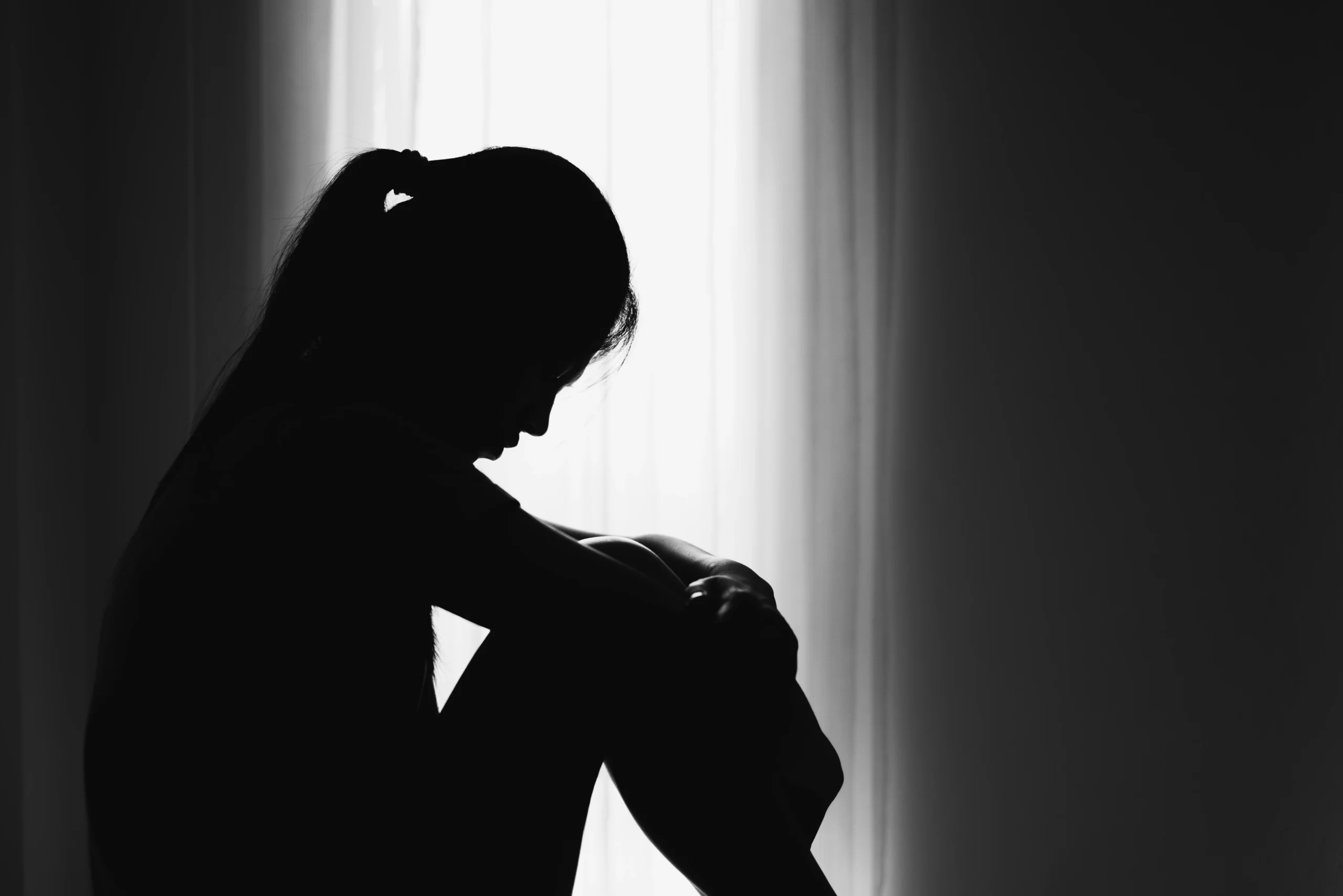 A sillhouette of a woman sitting in a room in front of a window with her arms around her knees
