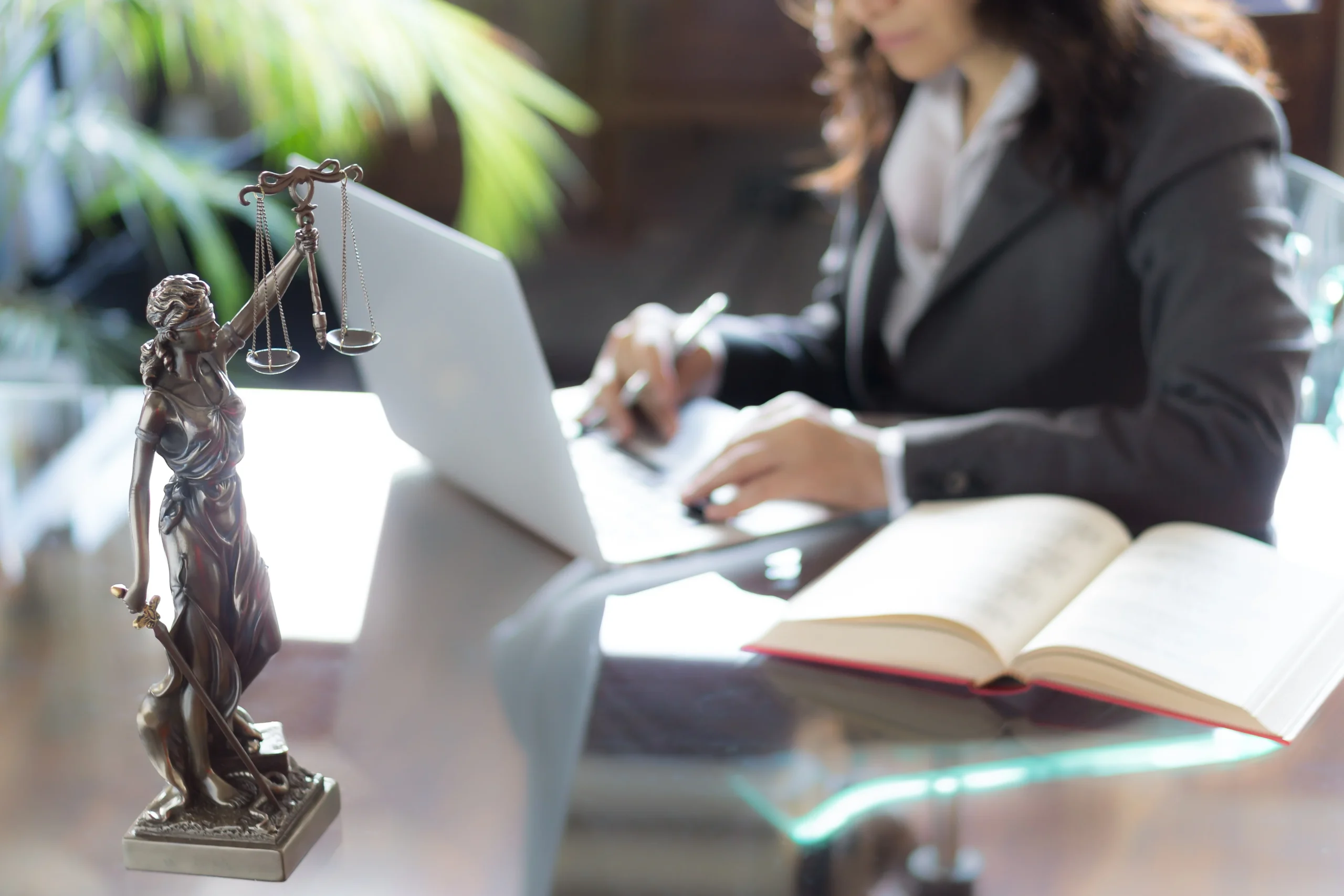 A business woman sits at a desk on a laptop with an open book next to her and a table-top statue of lady justice in front of her.