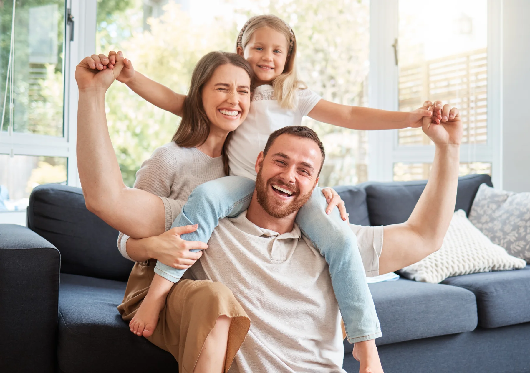 A mother sits on the couch, father sits on the floor in front of her, and their child sits on her father's shoulders.